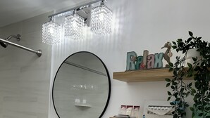 A bathroom with a white sink, a toilet, a shower with a glass-enclosed tub, a round mirror, and a shelf with toiletries. - Mariner Motel (Falmouth)