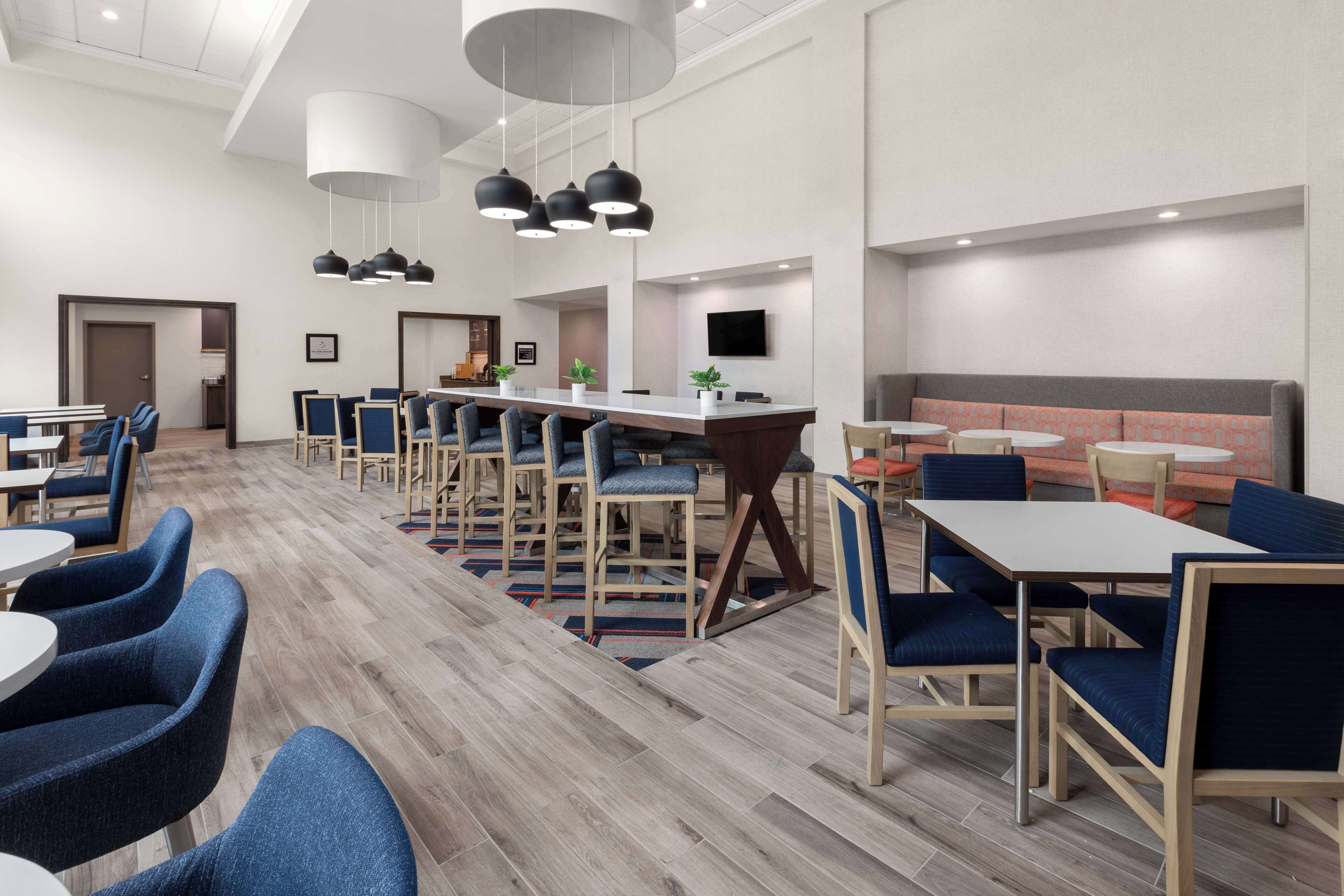 A modern hotel lobby with a long reception desk, multiple seating areas, and a large TV screen.
