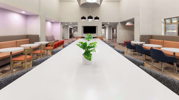 A spacious indoor area with a long white table, multiple seating options including orange and blue chairs, and a potted plant in the center.