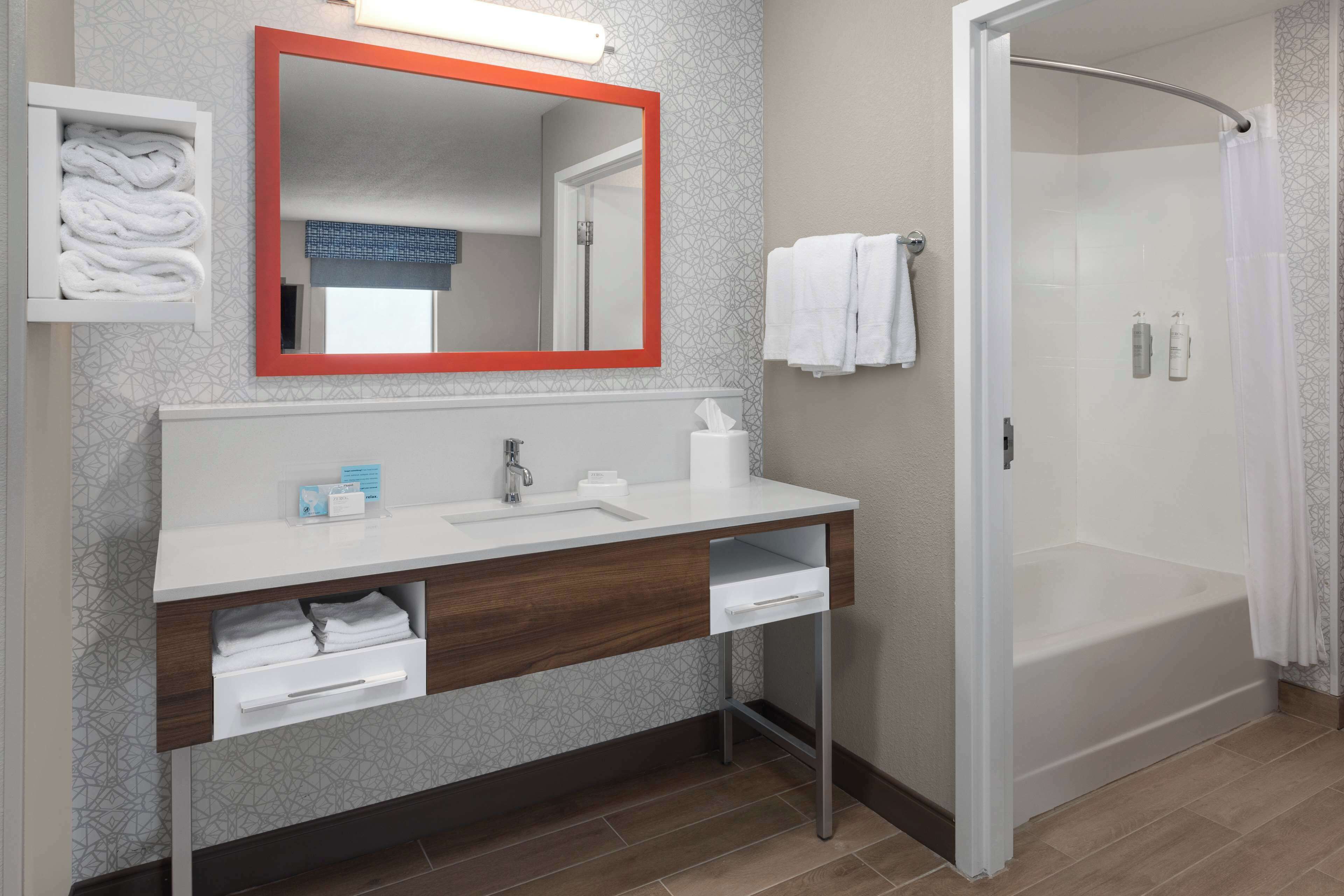 A modern bathroom with a white vanity, a large mirror with a red frame, a shower with a glass door, and neatly folded towels.