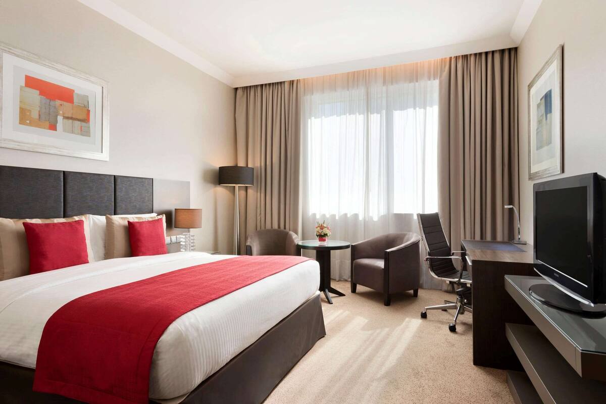 Ein Hotelzimmer mit einem großen Bett, einem Schreibtisch mit Computer, einem Sessel, einem Fernseher und einem Fenster mit Vorhängen.