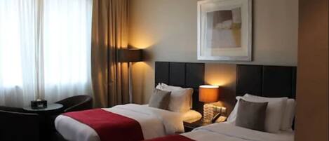 A hotel room with two beds, a desk, a chair, a lamp, and a framed picture on the wall.