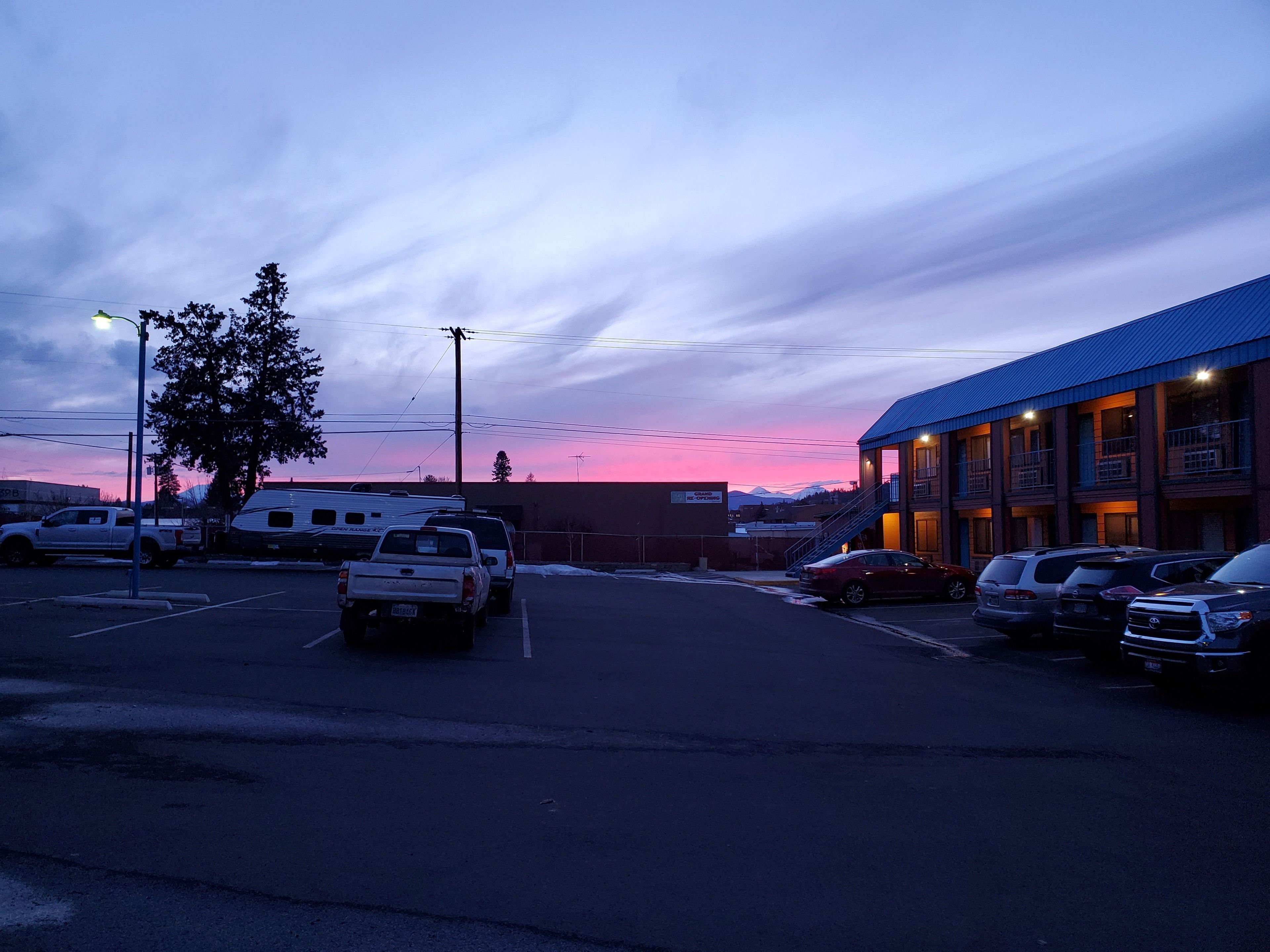 Un parking avec plusieurs véhicules, un bâtiment doté d’un toit bleu et un ciel au coucher du soleil.