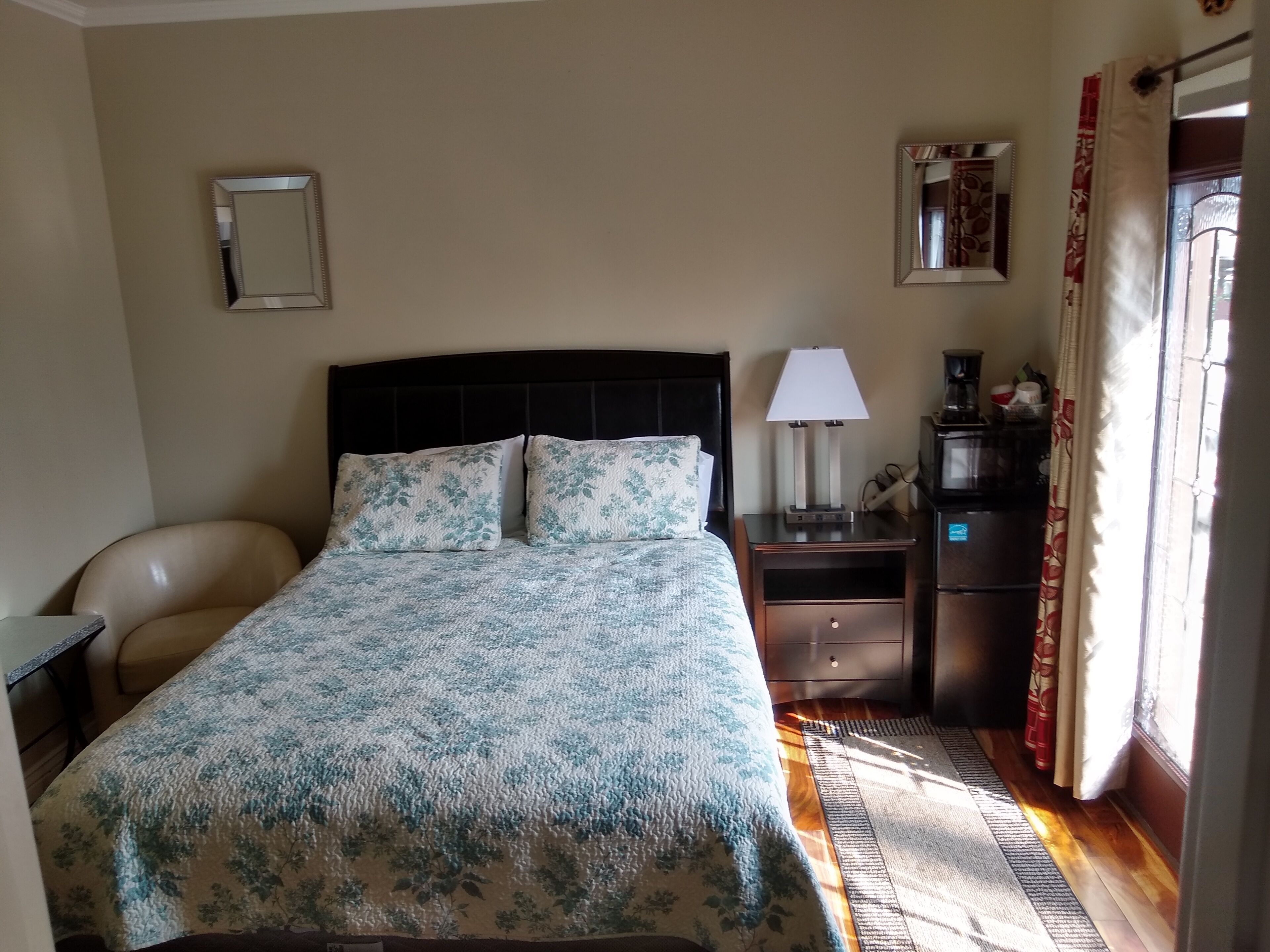 A neatly arranged hotel room with a bed, a chair, a nightstand with a lamp, a mirror, and a window with curtains.