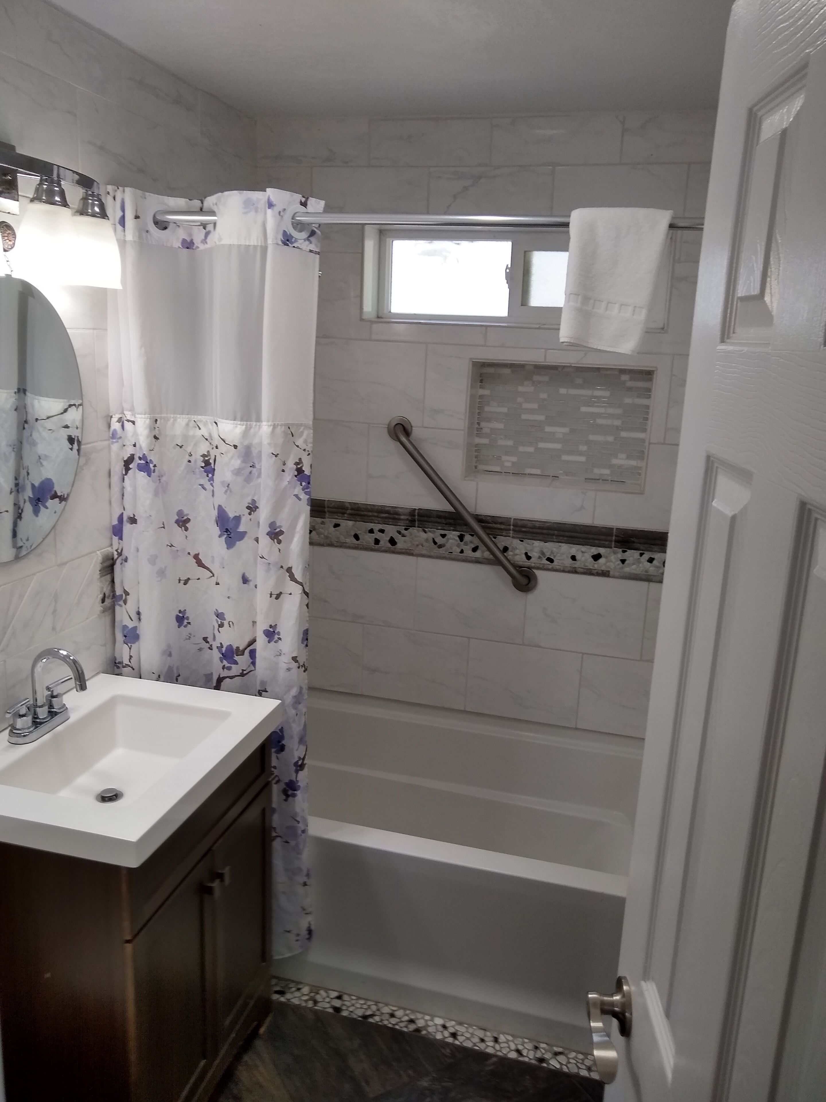 Un baño con un lavabo blanco, un espejo, ducha con barra de apoyo y una ventana sobre la ducha.