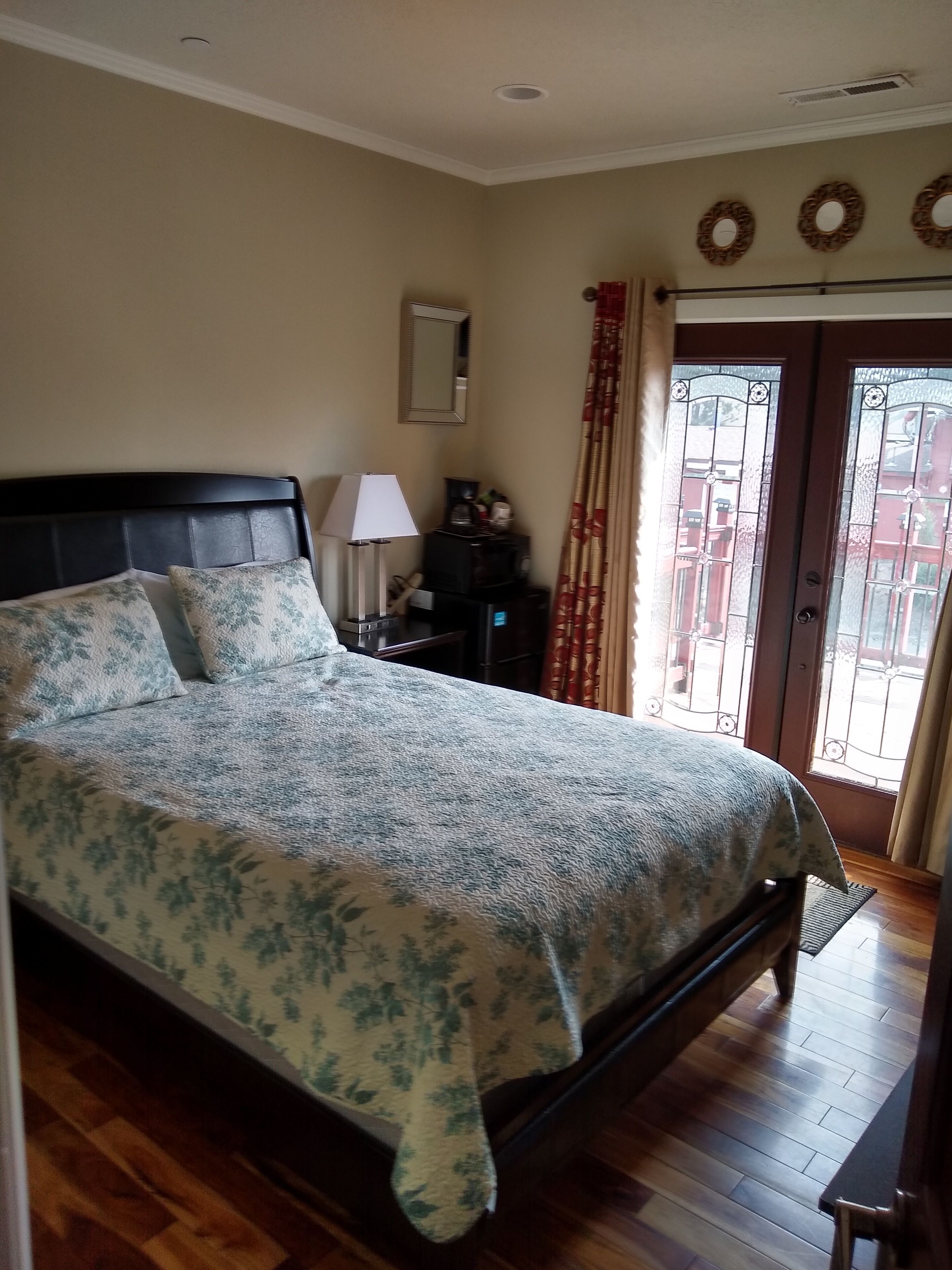 A bedroom with a large bed, a nightstand, a lamp, a mirror, and a view through a door with curtains.
