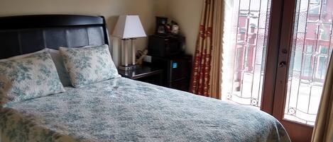 A bedroom with a large bed, a nightstand, a lamp, a mirror, and a view through a door with curtains.