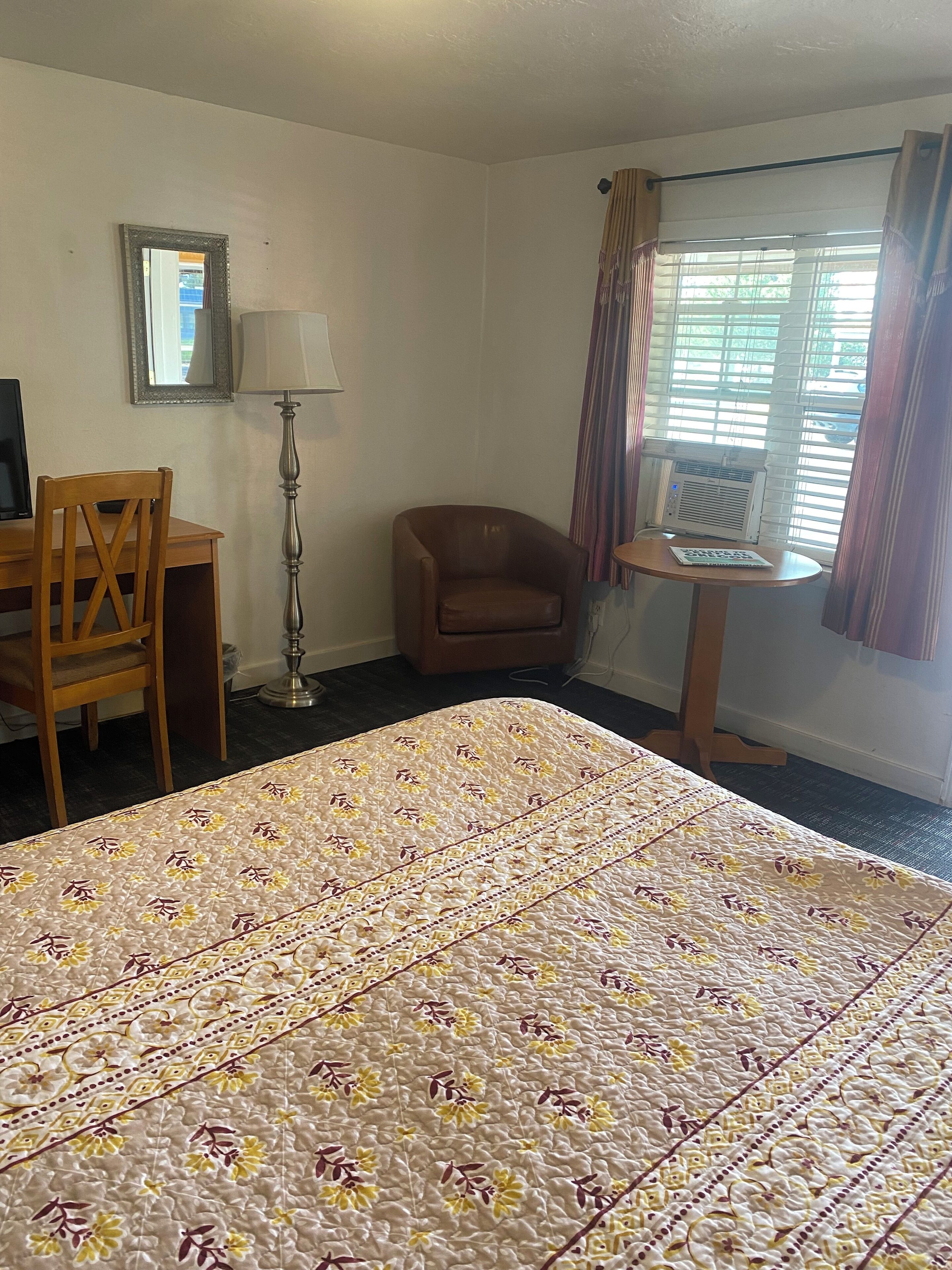 Une chambre d’hôtel avec un lit, un bureau, une chaise, une lampe, une petite table et une fenêtre avec des rideaux.