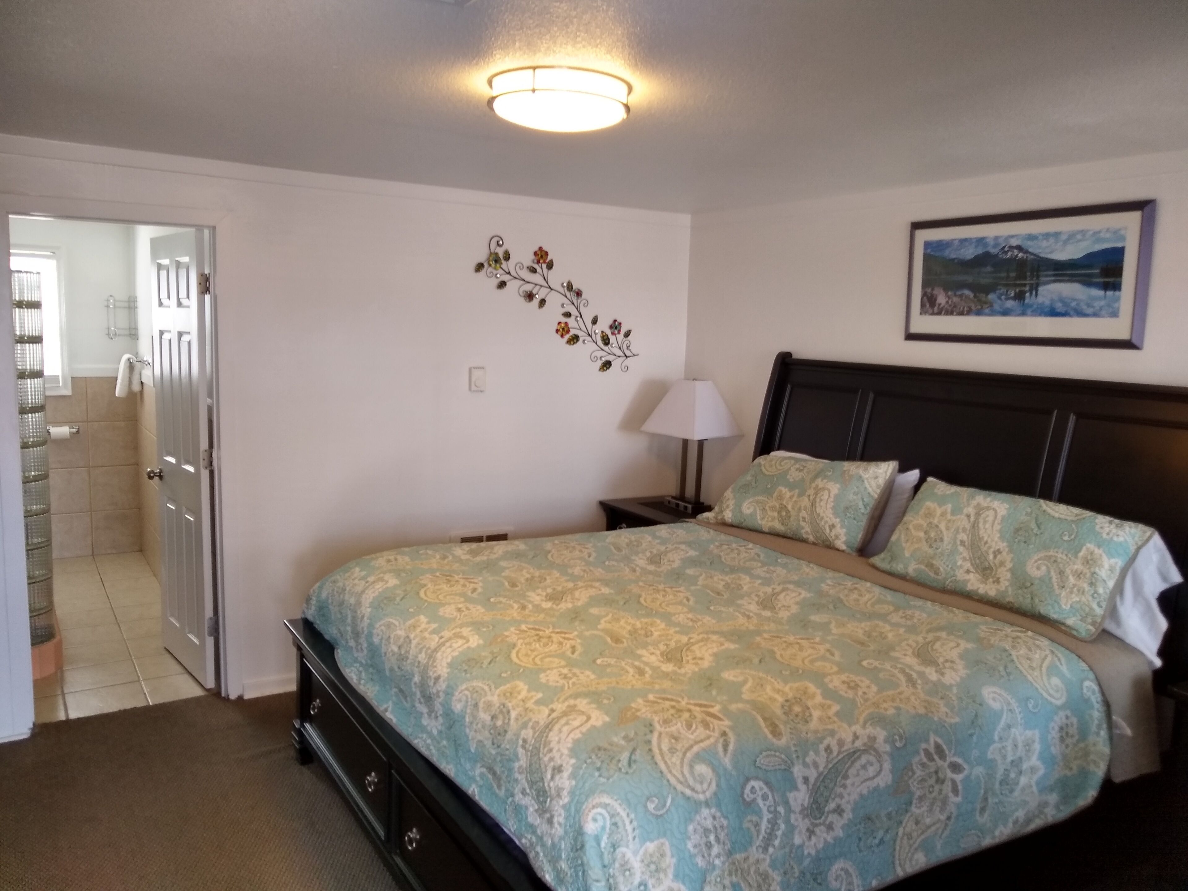 Un dormitorio con cama, una mesita de noche, una lámpara de pared, un adorno en la pared y un cuadro enmarco.