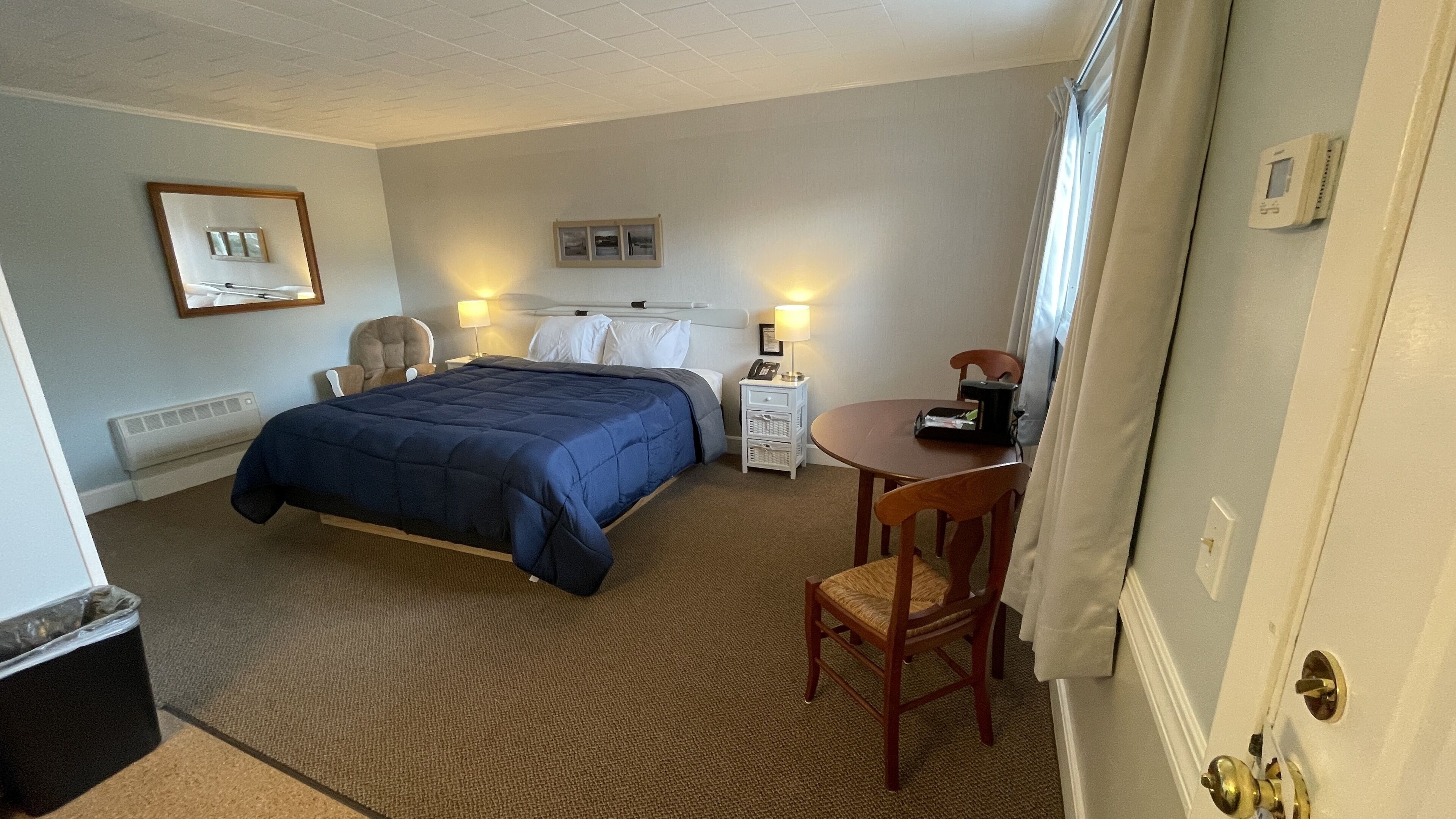 A hotel room with a bed, a chair, a small table, a nightstand, and a thermostat on the wall.