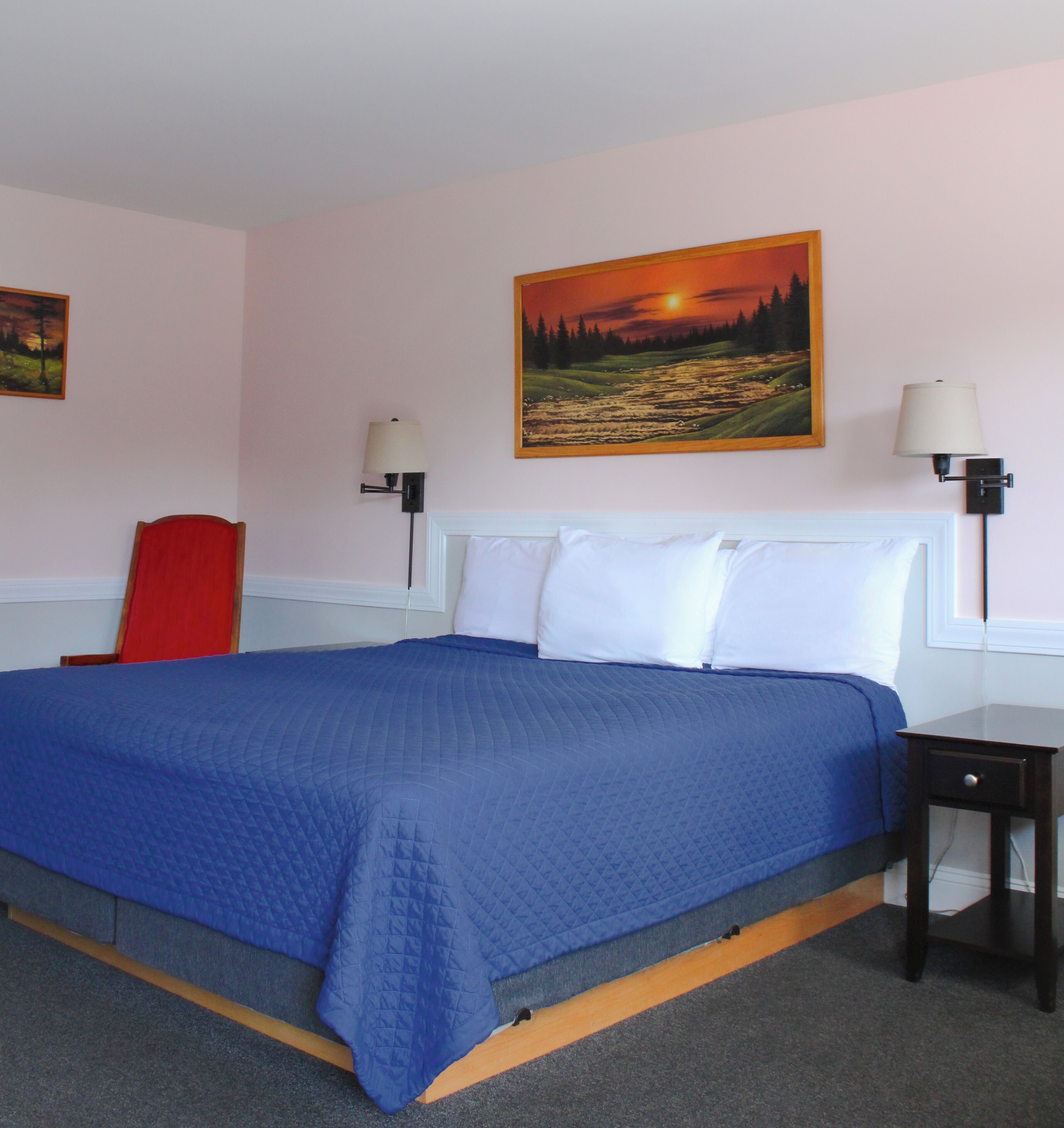 A hotel room with a large bed, a red chair, two wall-mounted lamps, and a painting of a sunset.