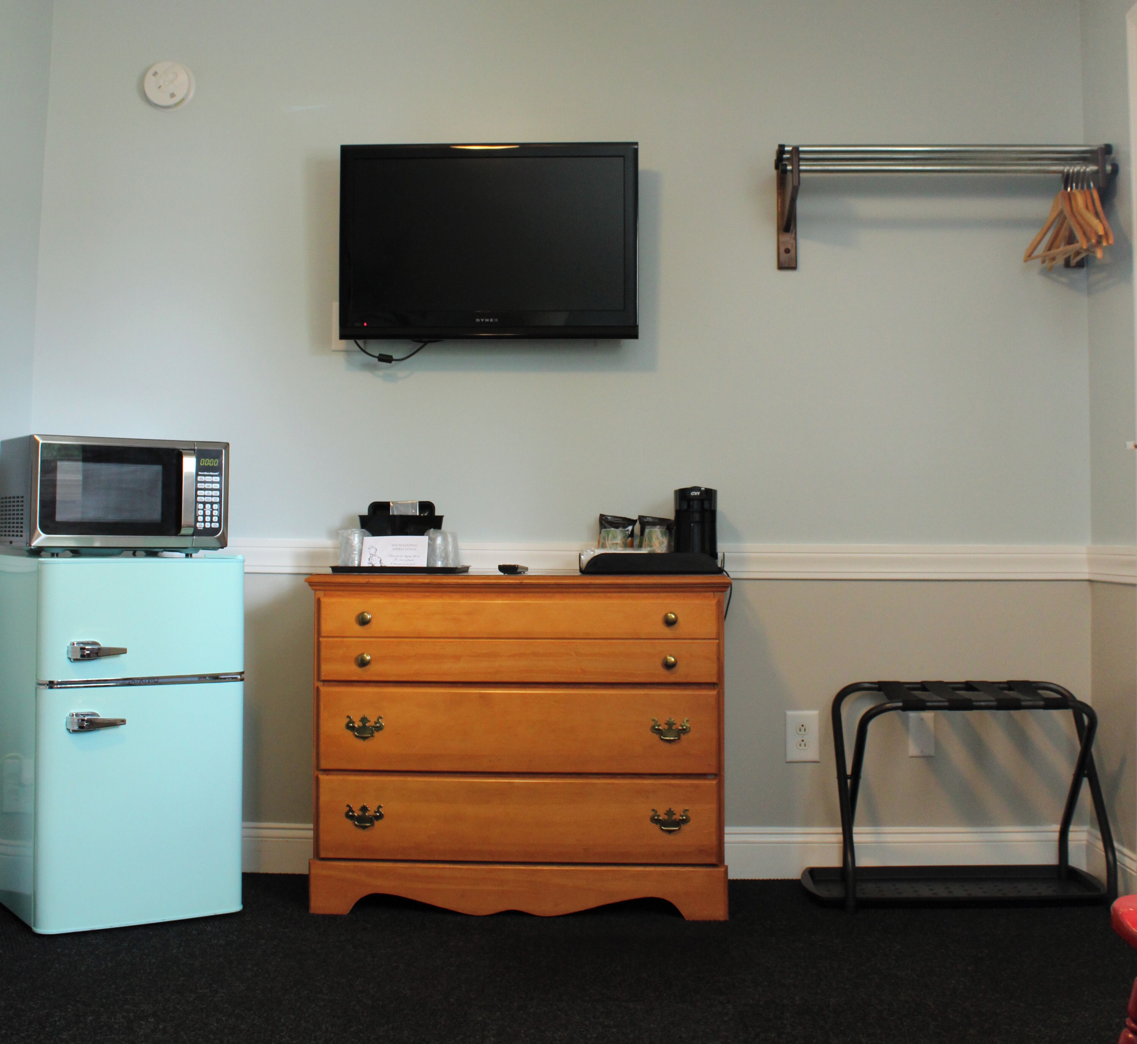 Een kamer met een lichtblauwe koelkast, een houten dressoir met lades, een magnetron, een zwarte koffiezetapparaat en een televisie aan de muur.