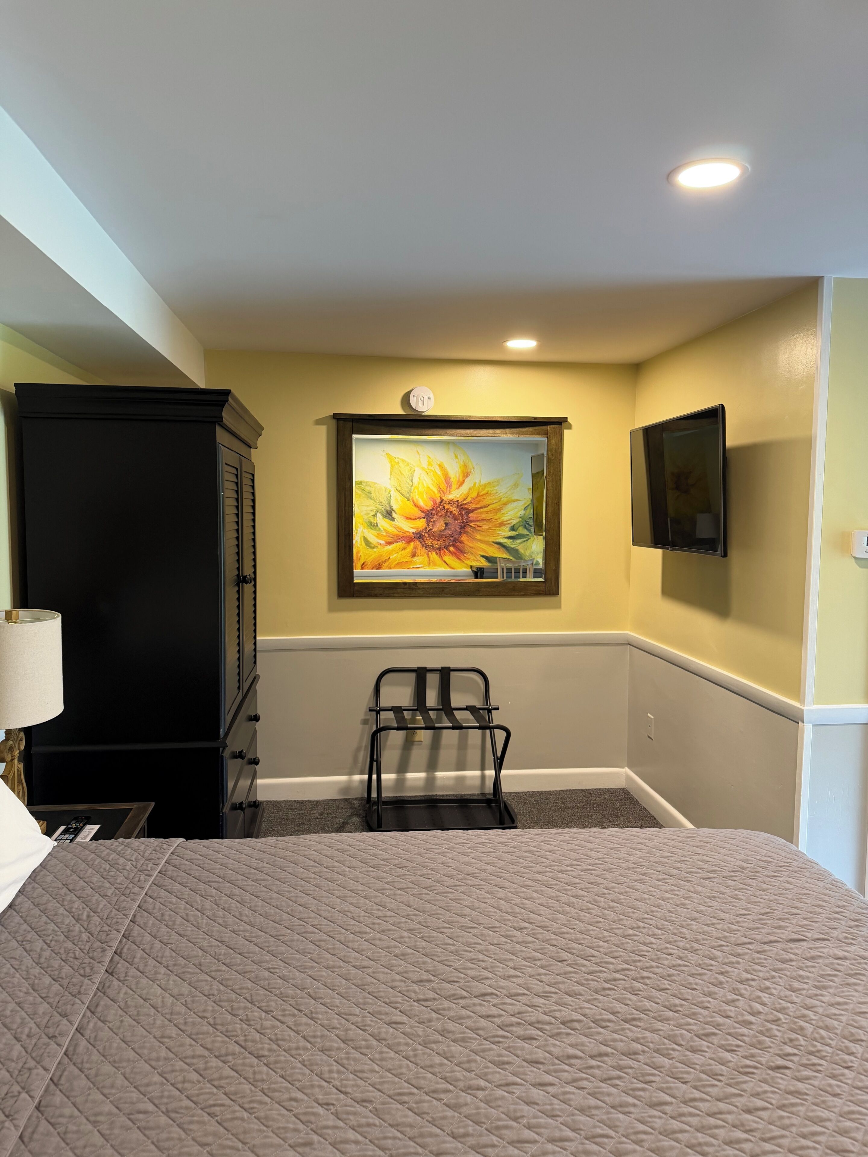 A hotel room with a bed, a painting of a sunflower, a TV mounted on the wall, and a black wardrobe.