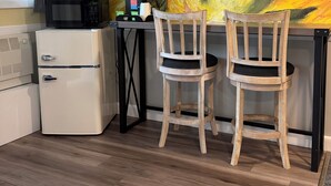 A kitchenette with a microwave, toaster, and coffee maker, two wooden chairs, and a wall with a floral painting.