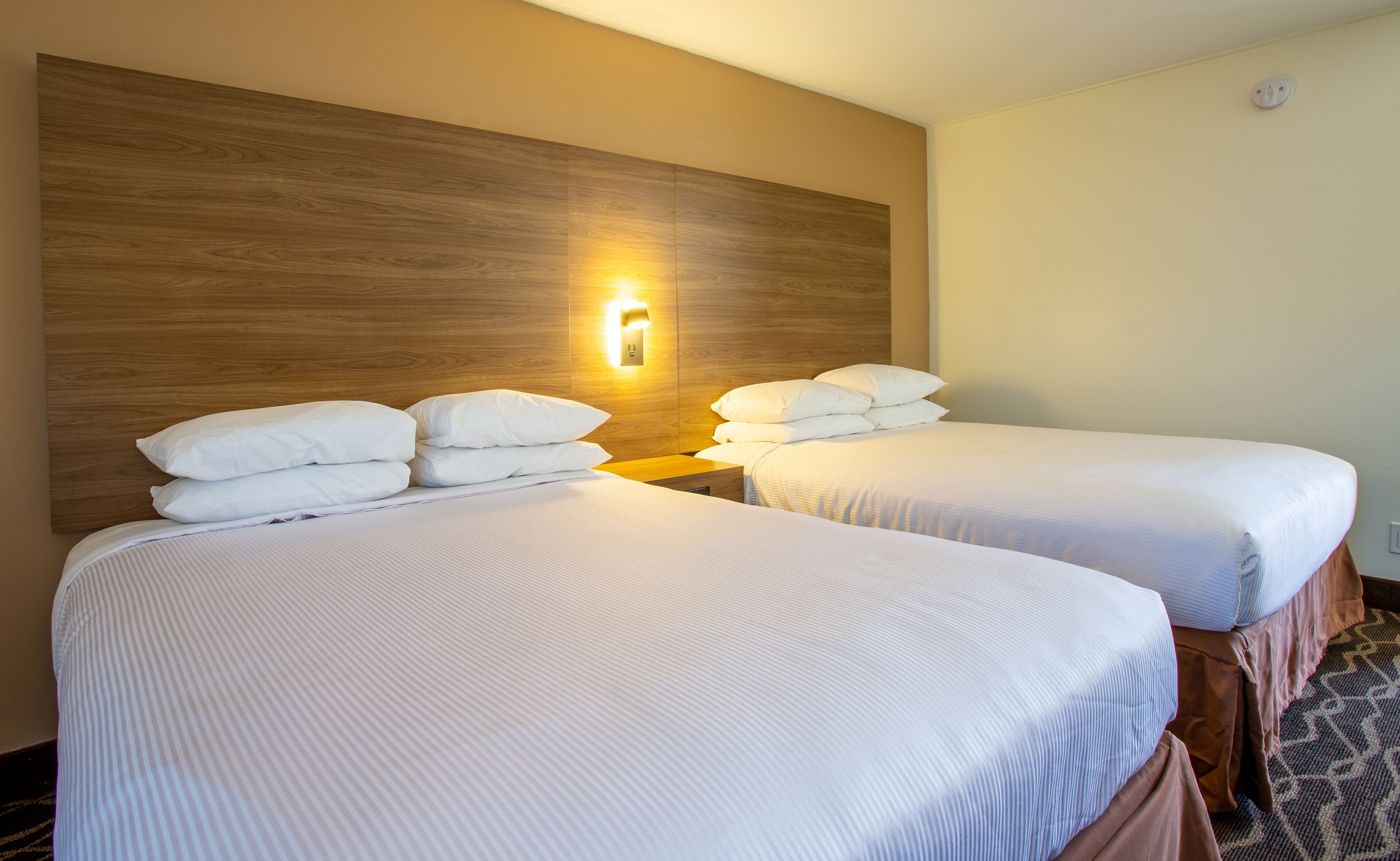 Two single beds with white linens and a wooden headboard, a wall-mounted light, and a smoke detector.