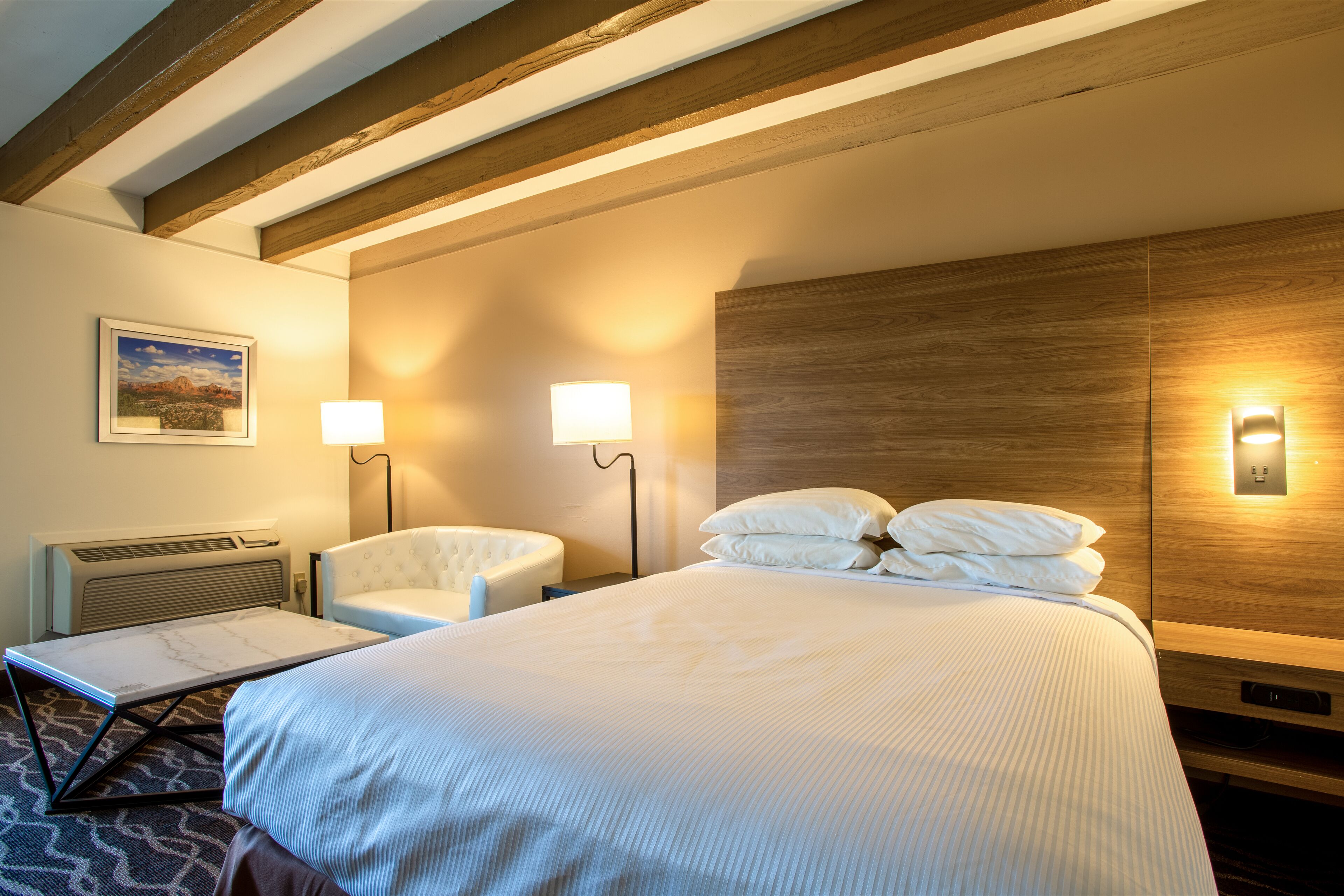 Un cuarto de hotel con una cama grande, dos lámparas de noche, una silla, una mesita y un cuadro en la pared.