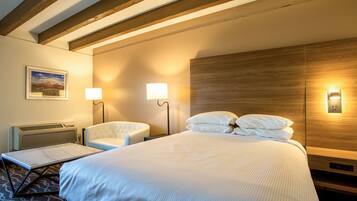 Un cuarto de hotel con una cama grande, dos lámparas de noche, una silla, una mesita y un cuadro en la pared.
