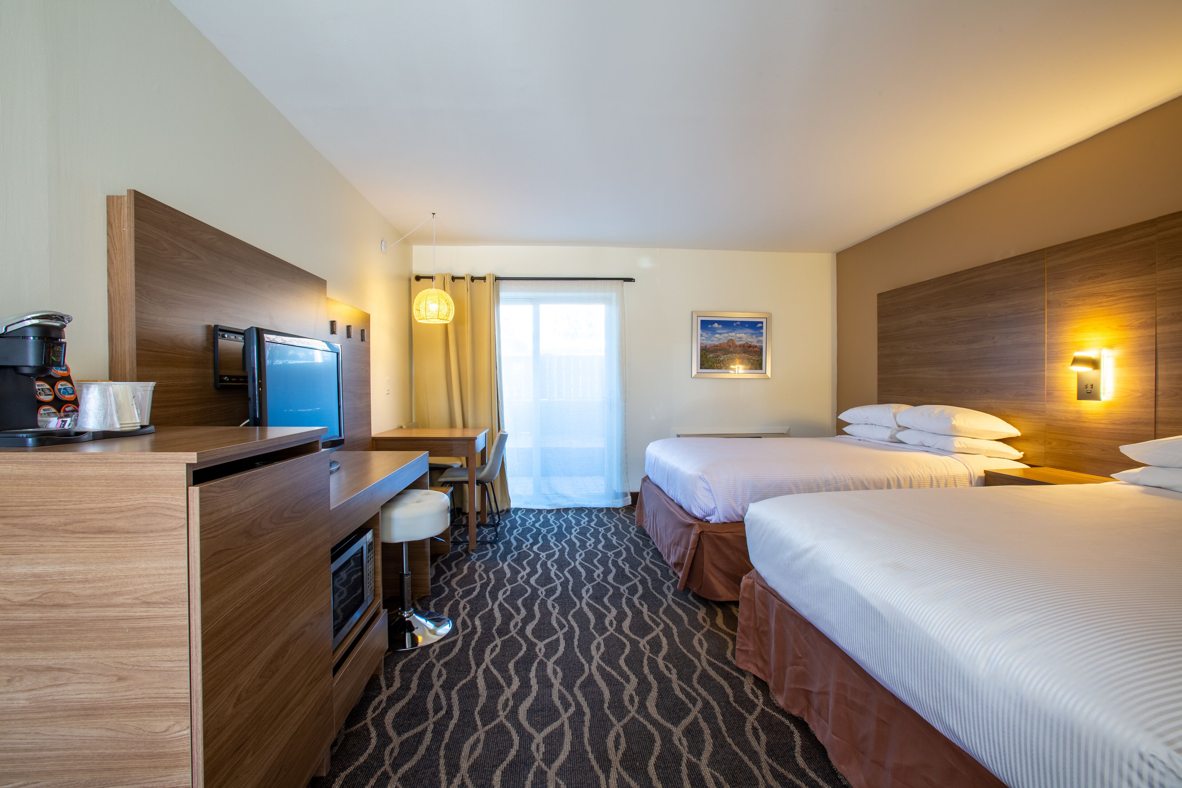 A hotel room with two beds, a desk with a computer, a coffee machine, and a view of the outdoors.