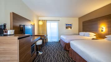 A hotel room with two beds, a desk with a computer, a coffee machine, and a view of the outdoors.