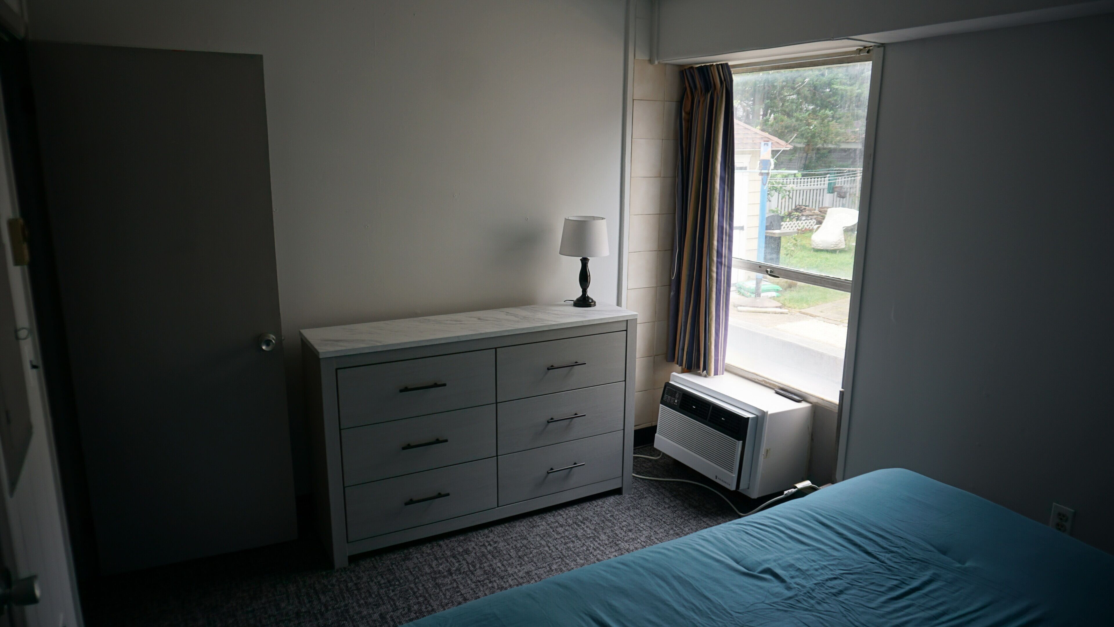 A bedroom with a bed, a dresser, a window with curtains, and a radiator.