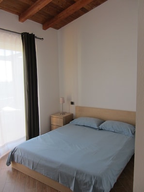 A bedroom with a wooden bed frame, a bedside table with a lamp, and a window with curtains.