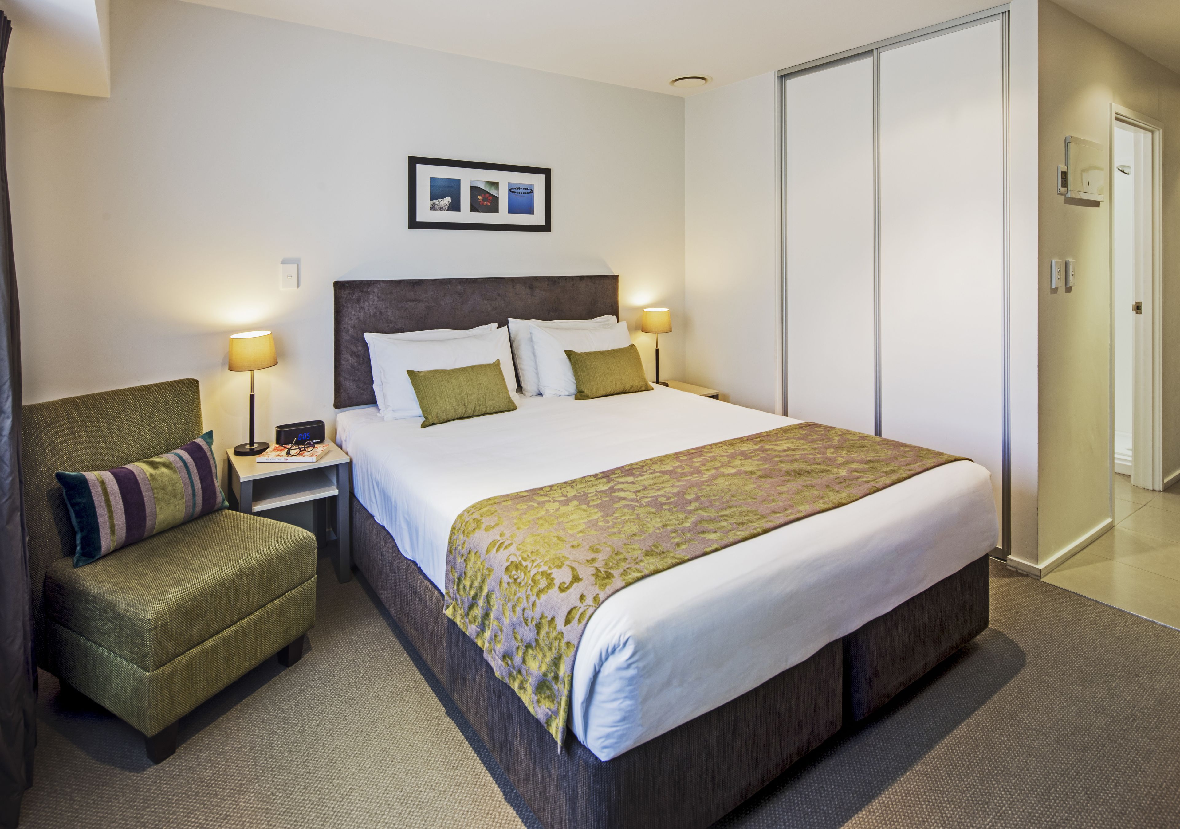A hotel room with a large bed, a chair, two bedside lamps, a framed picture on the wall, and a wardrobe.