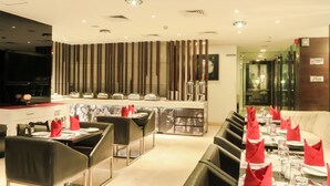 A modern restaurant with a long dining table set for multiple guests, a bar area, and a seating area with a cushioned bench. - The Nanee Suites (New Delhi)