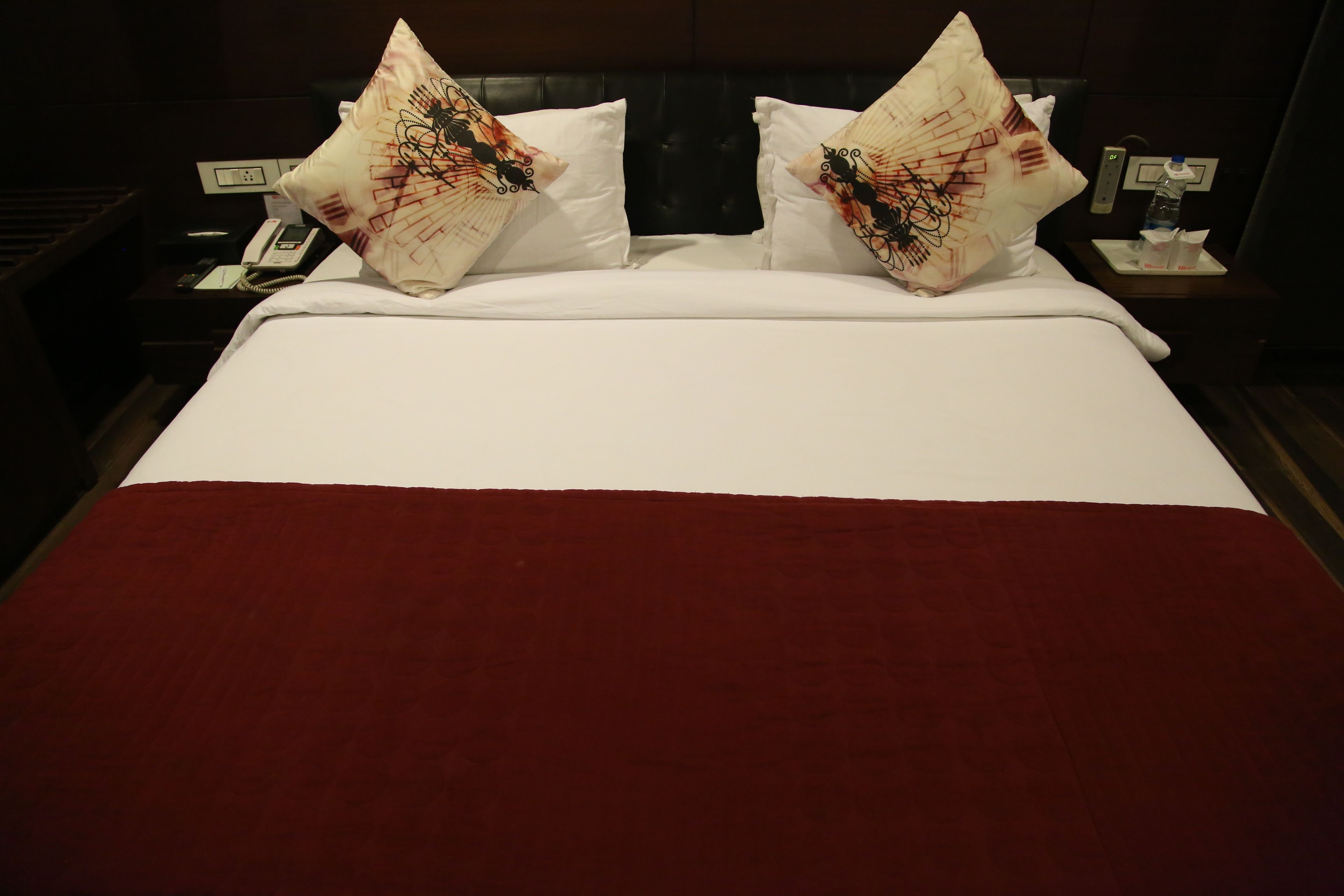 A hotel room with a large bed, two decorative pillows, a red throw blanket, a bedside table with a phone, and a nightstand with a lamp.