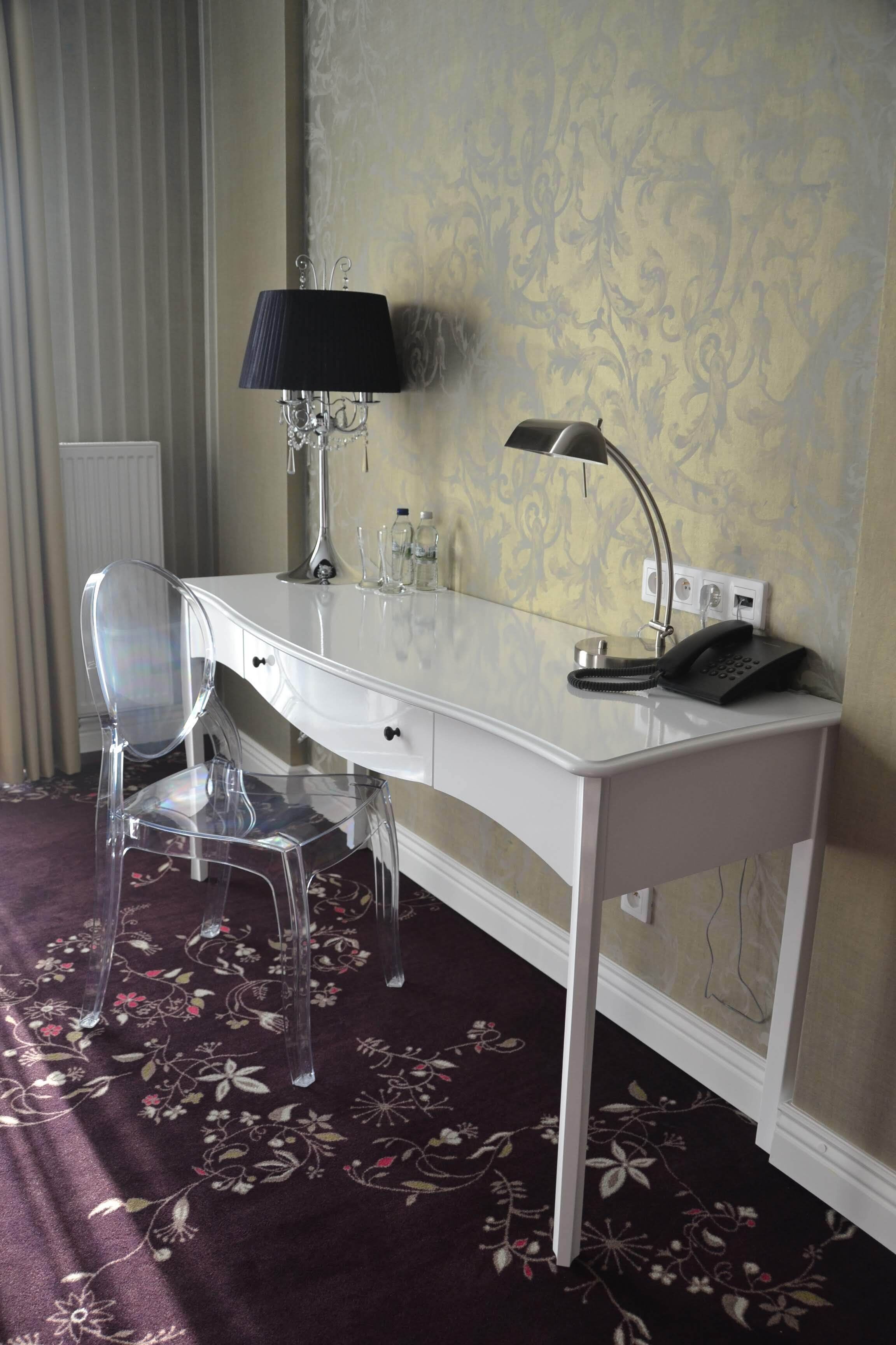Une chambre d’hôtel avec un bureau blanc, une chaise transparente, une lampe, un téléphone et un radiateur.