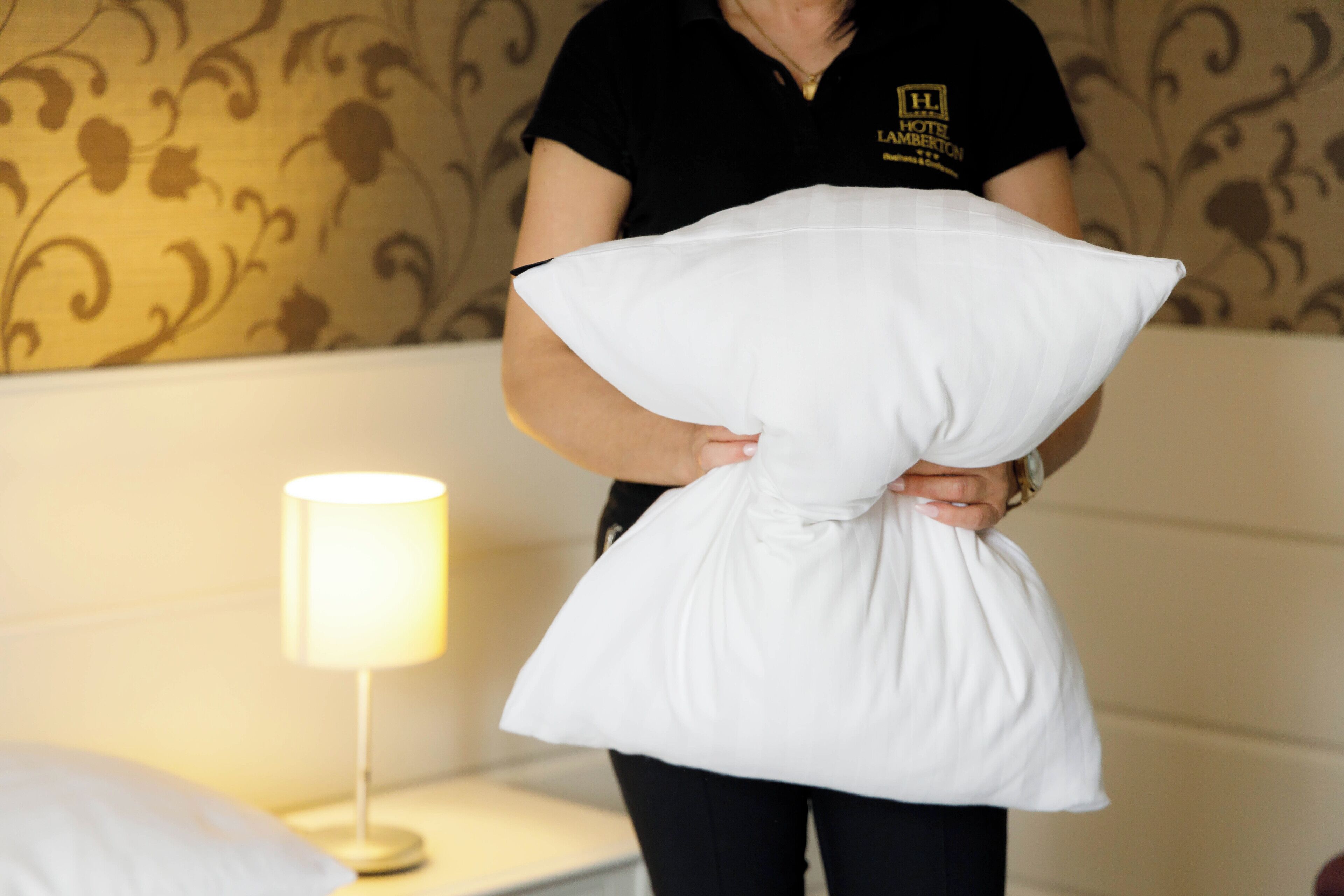 Una persona sosteniendo un almohadón en una habitación con papel tapiz estampado y una lámpara de noche.