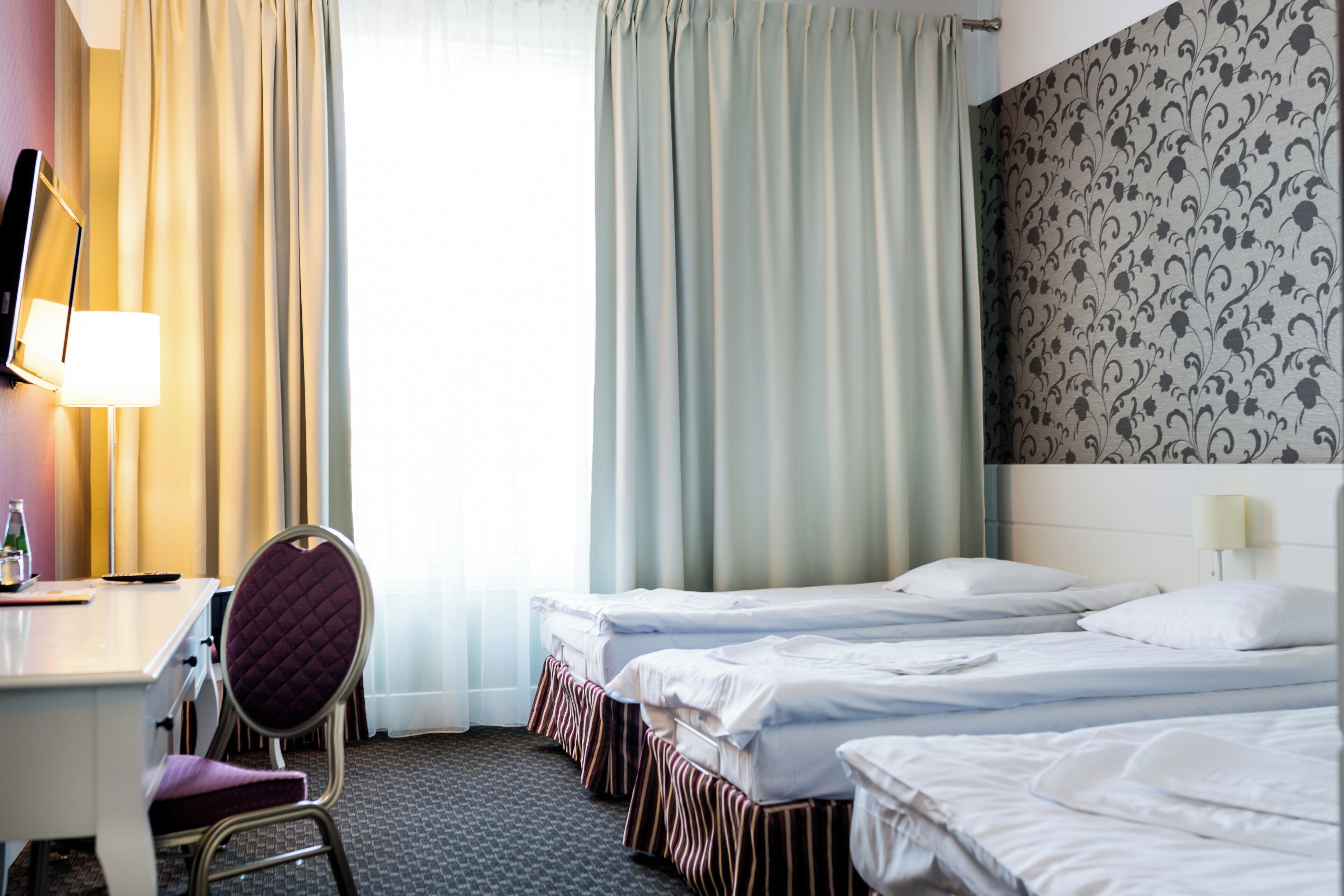 A hotel room with a desk, two beds, a chair, and a large window with curtains.