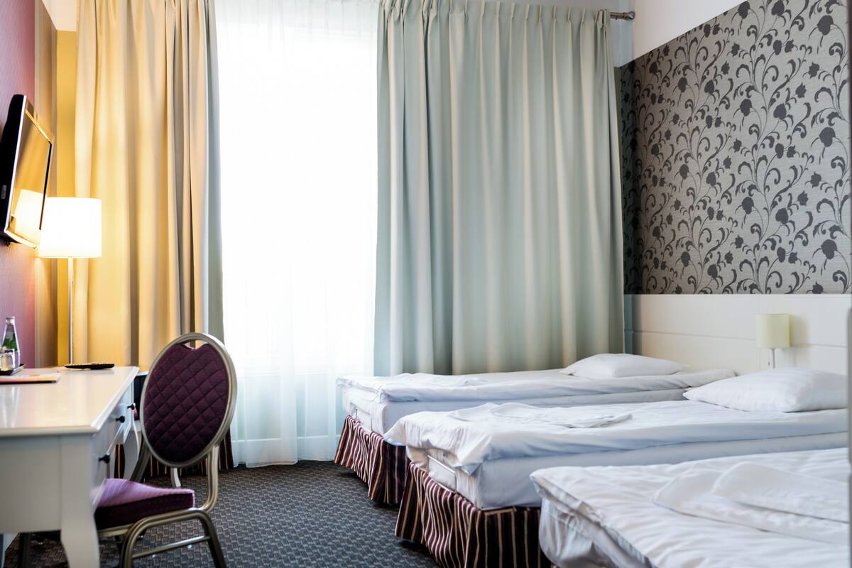 A hotel room with a desk, two beds, a chair, and a large window with curtains.