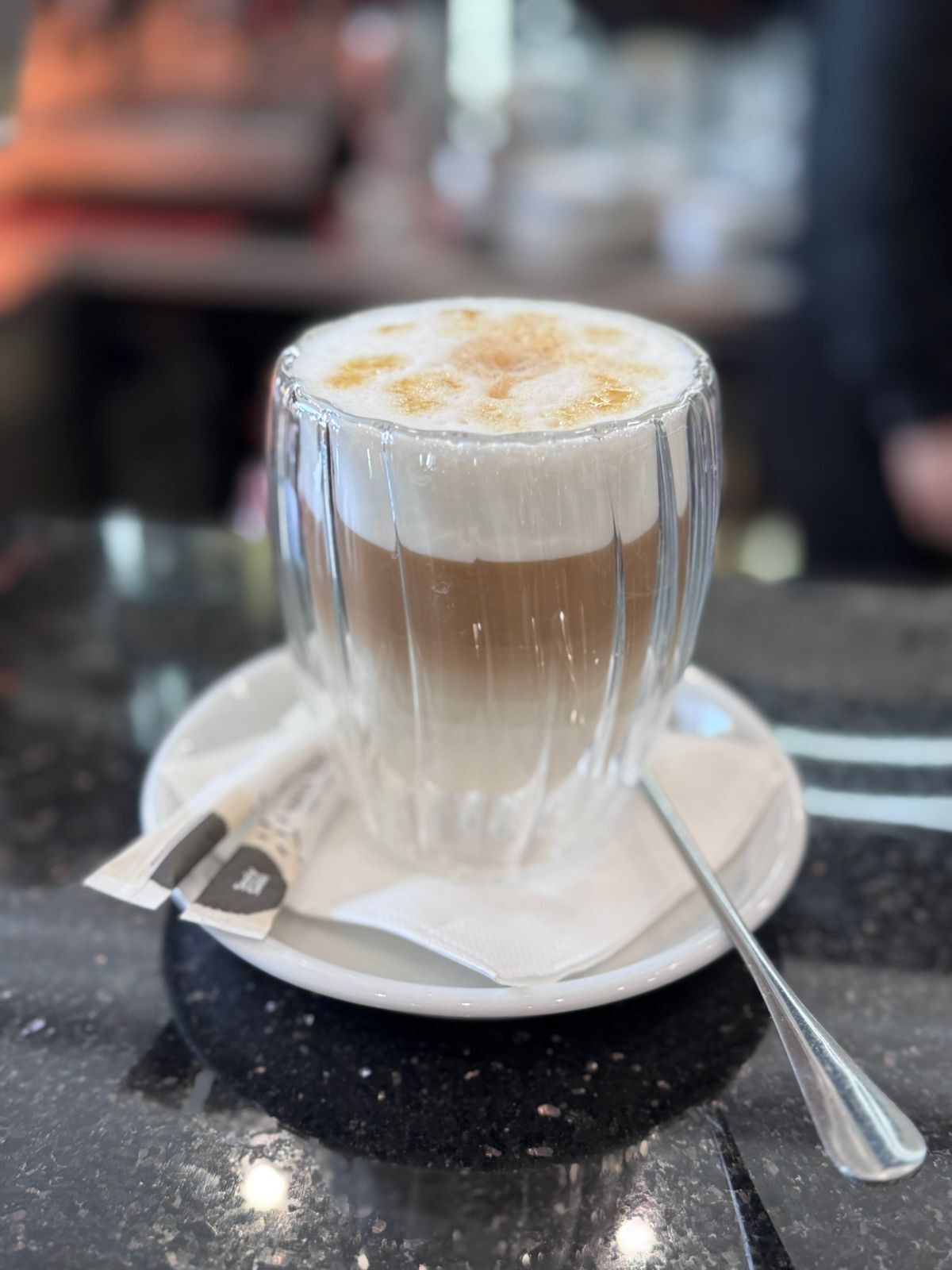 A clear glass cup of coffee with a spoon on a saucer, placed on a marble surface.
