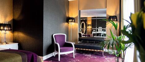 A hotel room with a bed, a purple armchair, a sofa, a large mirror, and a potted plant.