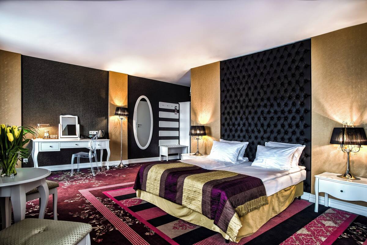 A bedroom with a bed, bedside tables, a desk, a chair, a mirror, and a potted plant.