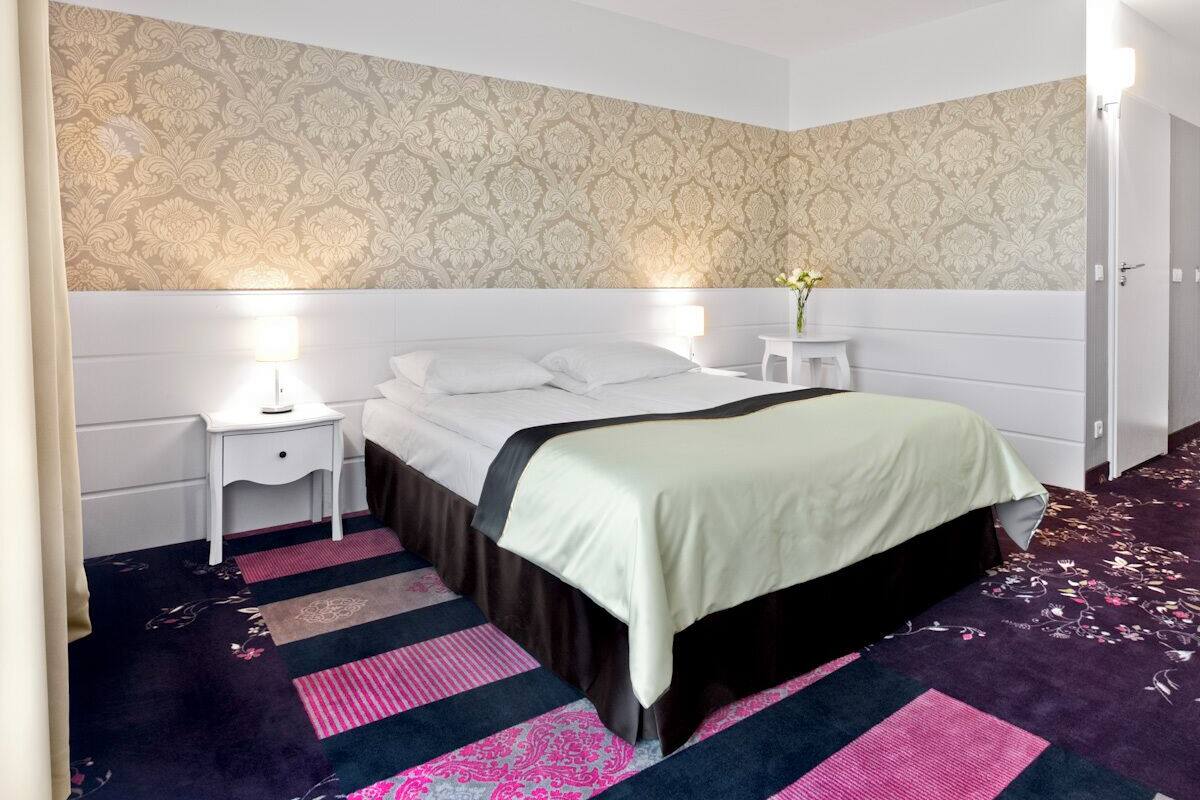 A bedroom with a bed, nightstands, a lamp, a vase with flowers, and a patterned carpet.