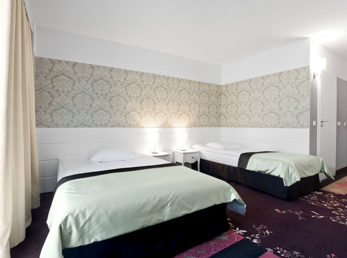 A hotel room with two beds, a patterned wallpaper, a nightstand with a lamp, and a door.