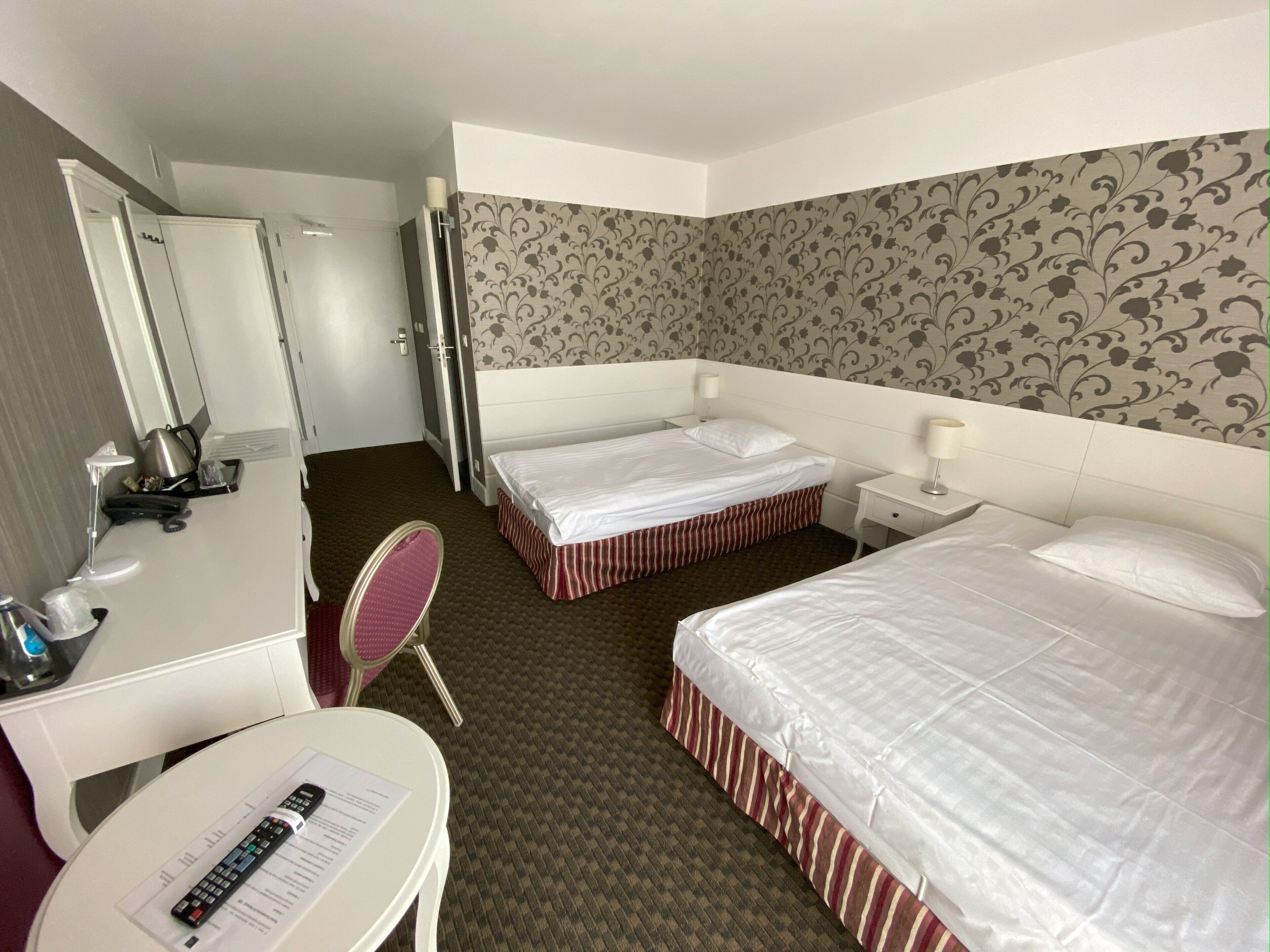 A hotel room with two beds, a desk with a chair, a mirror, a small table with a lamp, and a patterned wall.