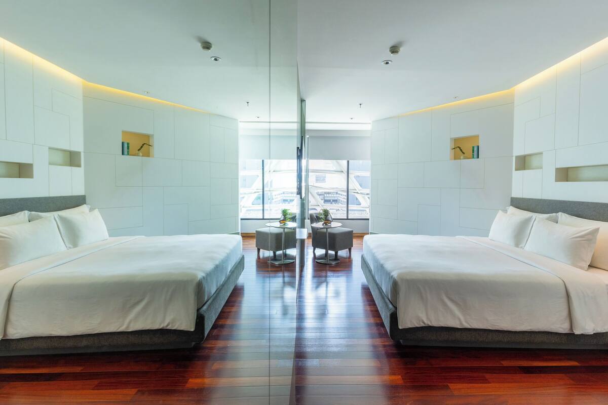 A mirrored hotel room with two beds, a central table, and a view of the city through large windows.