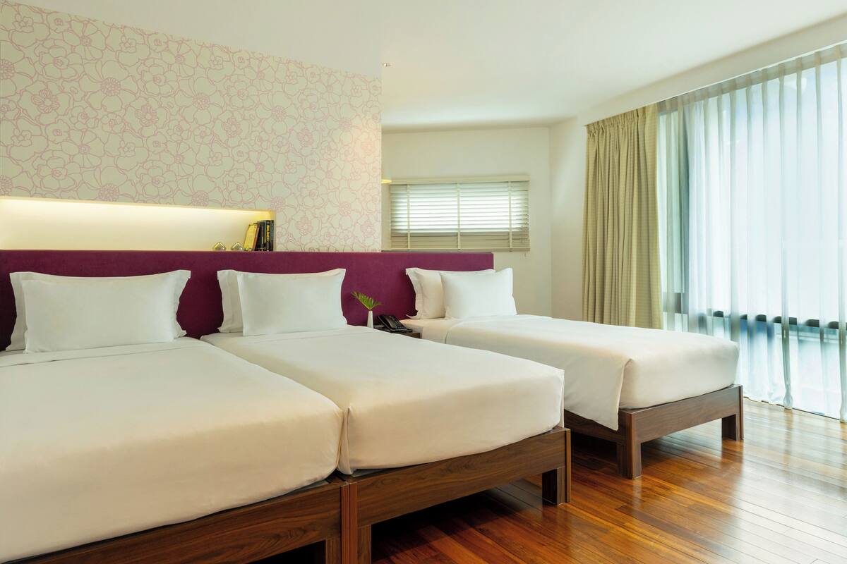 A hotel room with two beds, a wooden floor, a floral wallpaper, and a large window with curtains.
