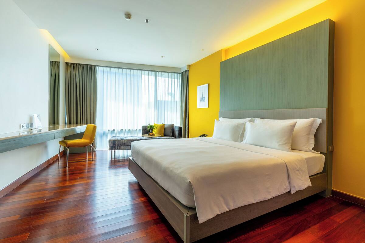A modern hotel room with a large bed, a seating area with a chair and a small table, and a large window with curtains.