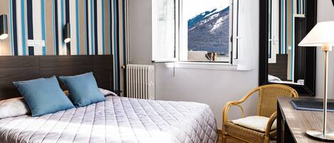 A neatly made bed with a quilted blanket, a wicker chair, a desk with a lamp, and a window with a mountain view.