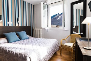 Un letto rifatto con coperta trapuntata, una sedia in vimini, una scrivania con una lampada e una finestra con vista sulle montagne.