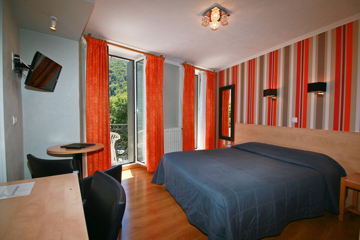 A bedroom with a bed, a desk, and a chair. There is a window with curtains, a TV mounted on the wall, and a ceiling light fixture.