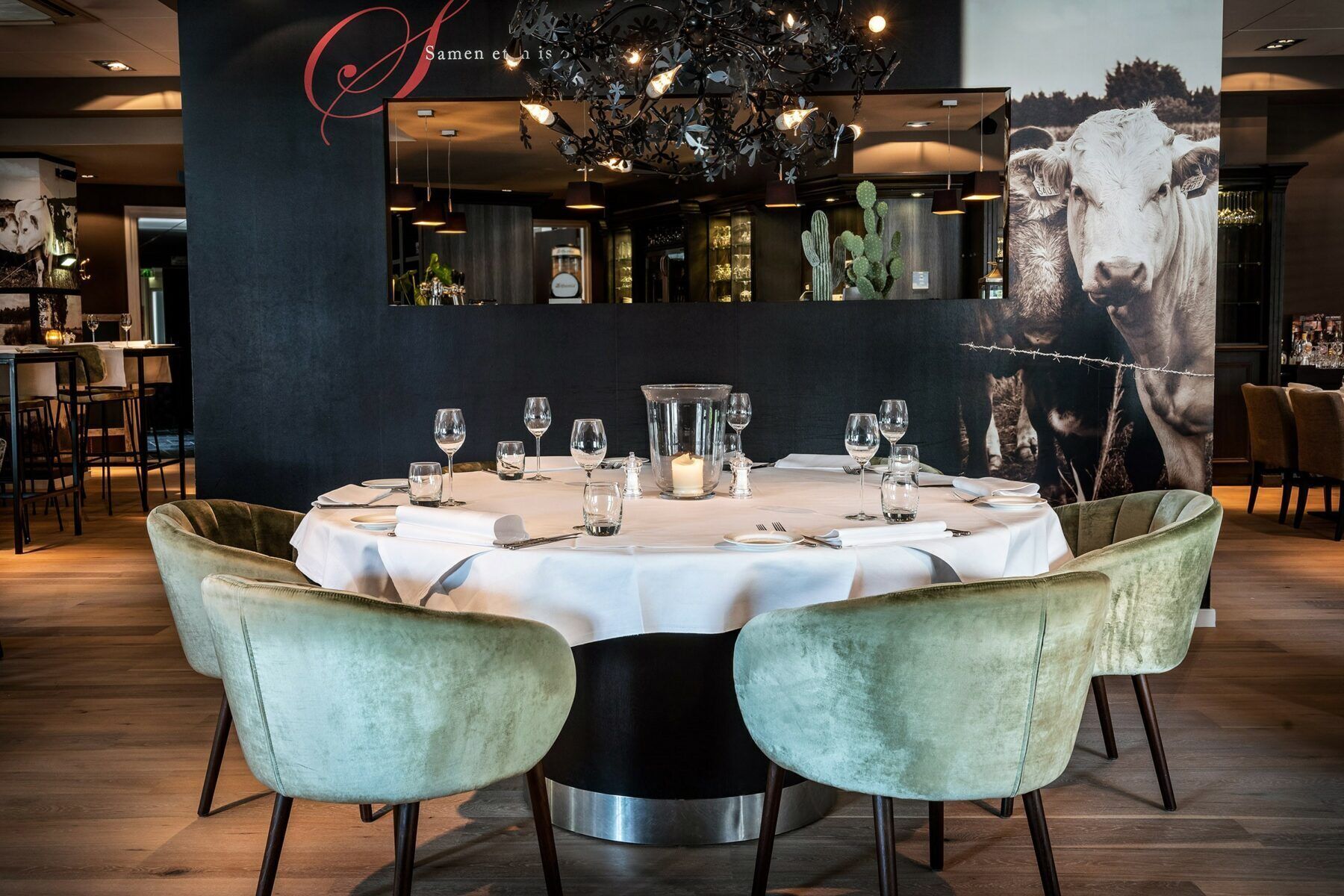 Un restaurante con una mesa redonda lista para comer, dos sillas de terciopelo, una araña y un mural grande en la pared.