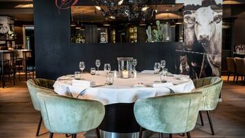 Un restaurante con una mesa redonda lista para comer, dos sillas de terciopelo, una araña y un mural grande en la pared.