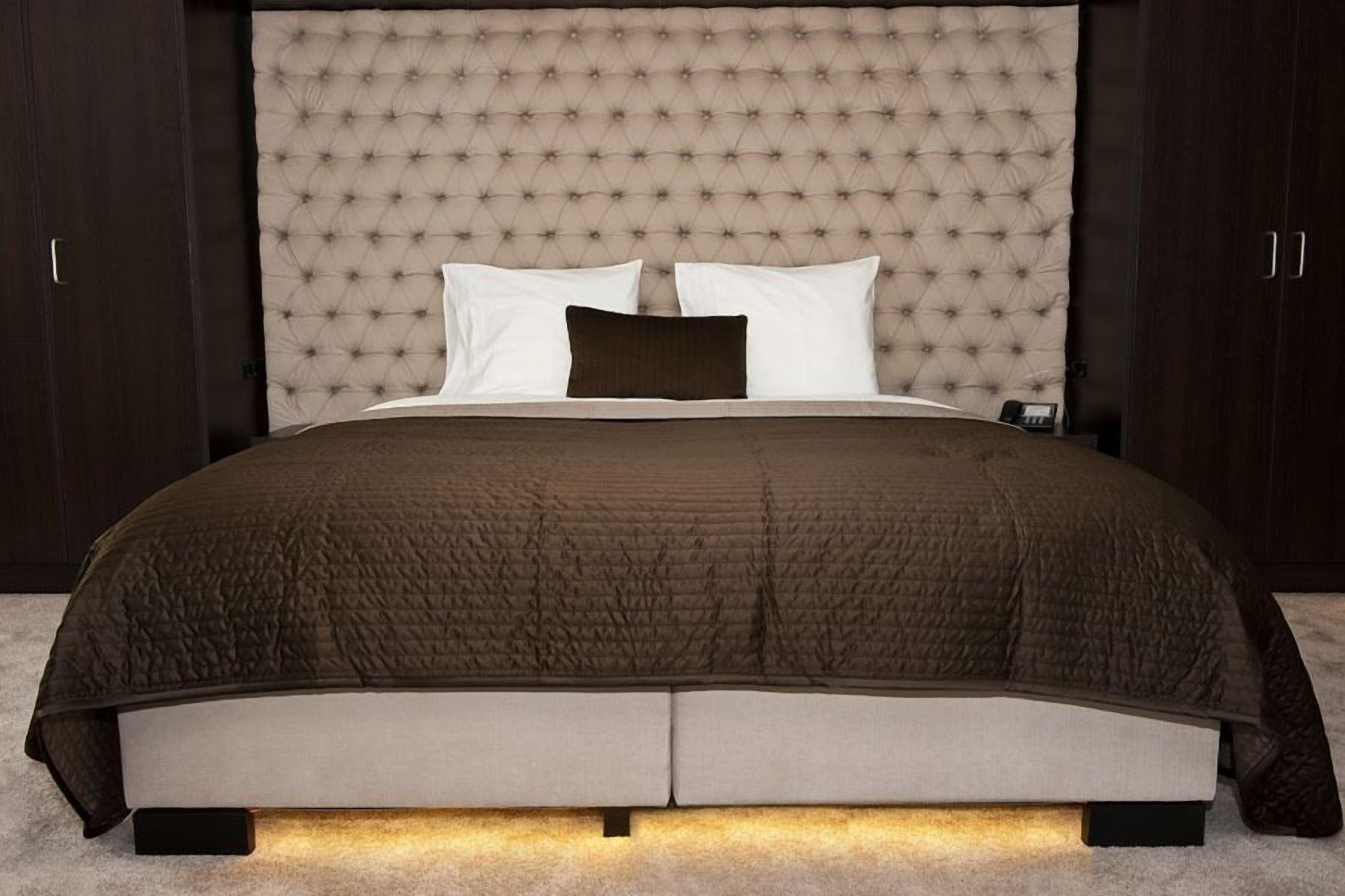 Un dormitorio con cabecera capitonada, cama con colcha acolchada oscura y mesita de noche con lámpara.