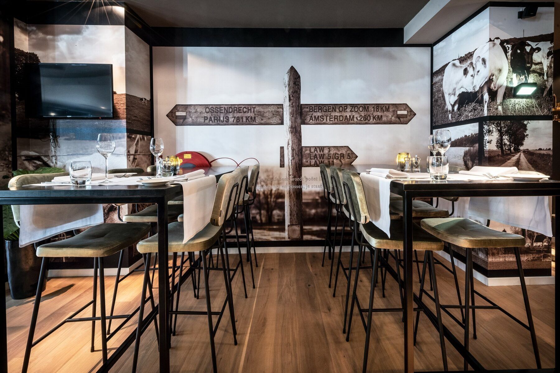 A restaurant with a central wooden signpost indicating distances to various destinations. Tables are set with glasses and cutlery, and the room has a modern, rustic design with wooden flooring and metal chairs.