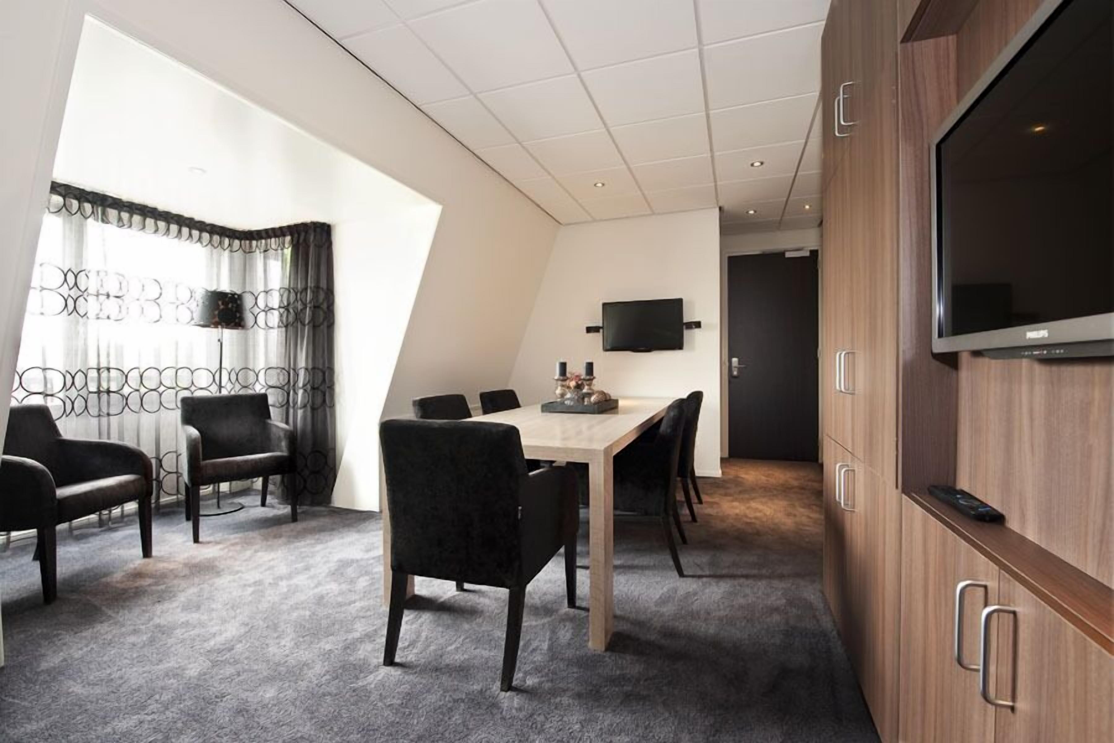 Una habitación de hotel moderna con comedor, televisión de pantalla plana y un ventanal con cortinas.
