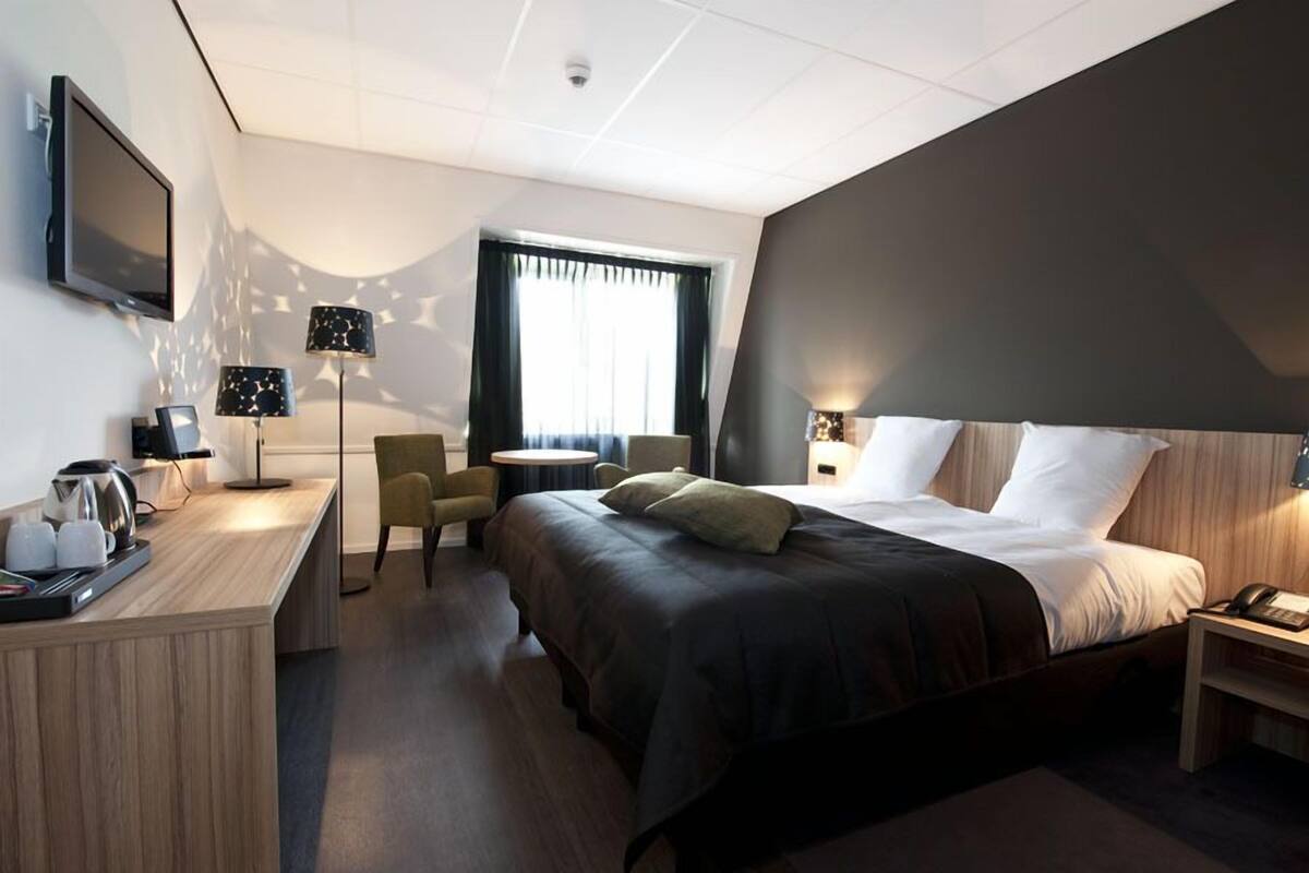 A modern hotel room with a large bed, a desk with a coffee maker, a TV, and a seating area with chairs and a table.